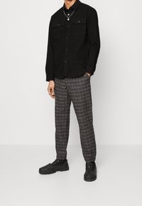 Schwarzes Hemd mit Knopfleiste und zwei Brusttaschen, kombiniert mit braunen karrierten Hosen. Dazu schwarze klobige Schnürschuhe.