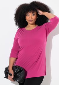 Top lungo fucsia con maniche lunghe, scollo rotondo e spacchi laterali, abbinato a una borsa nera arricciata. Tessuto morbido e leggero.