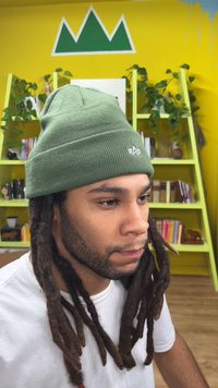 Gorro de malha verde com textura canelada e um logótipo na frente. Cabelo com dreadlocks e uma camisa branca são visíveis contra um fundo amarelo brilhante.