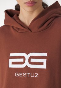 Osoba oblečená v hnědé mikině s bílým stylizovaným logem "GG" a textem "GESTUZ" vyšívaným na hrudi.