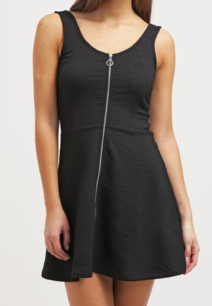 Femme portant une robe noire texturée sans manches avec une fermeture éclair argentée à l'avant et une jupe évasée, sur un fond neutre.