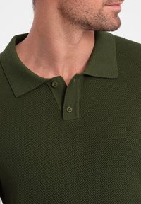 Tmavě zelené polo tričko vyrobené z texturované látky; má límec a dvě knoflíkové zapínání. Žebrovaná textura na těle.