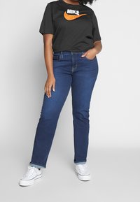 Černé tričko s bílým logem Nike a oranžovým swoosh, spárováno s modrými rovnými džíny, které jsou ohrnuté na manžetách, a bílými teniskami.