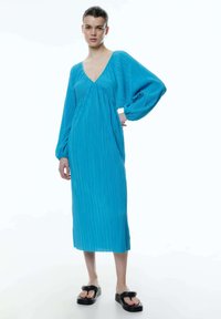 Blauwe plooienjurk met een diepe V-hals en lange, wijd uitlopende mouwen. Midi-lengte met een vloeiende silhouet. Gemaakt met zwarte platformsandalen.