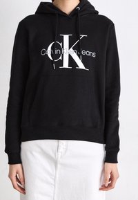 Černá mikina s kapucí z měkké látky s velkým bílým logem "CK" a nápisem "Calvin Klein Jeans". Obsahuje klokaní kapsu a stahovací šňůrky.