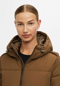 Veste matelassée marron avec fermeture éclair, capuche réglable et deux poches latérales. Le tissu a une texture lisse et une finition mate.