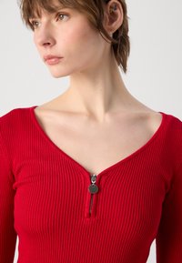T-shirt long rouge à côtes, à manches longues, doté d'un décolleté en V profond et d'une fermeture éclair à l'avant avec un tirette circulaire. Texture lisse et silhouette ajustée.