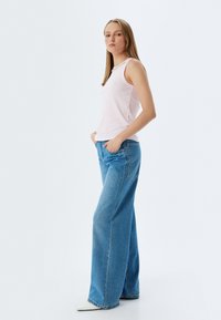 Hellrosa, gerippte Tanktop mit einem runden Ausschnitt, kombiniert mit weit geschnittenen, hellblauen Jeans und spitzen weißen Schuhen, die Hände in den Taschen.