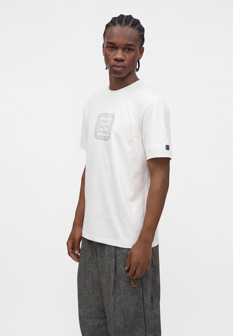 Young man with braided hair wearing a white T-shirt with a geometric logo and loose gray pants, standing against a plain white background.