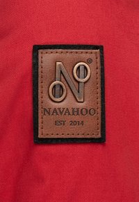 Bruine leren label met een reliëf logo "N", omringd door zwarte stof, vastgestikt op rode materiaa. De tekst luidt "NAVAHOO EST 2014".