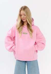 Felpa oversized rosa realizzata in tessuto morbido, con tasca frontale e logo ricamato verde. I polsini e l'orlo sono rifiniti con cuciture lisce.