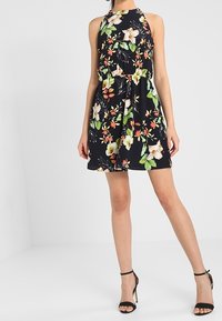 Svart blommig klänning med halterneck, prydd med vita blommor och gröna blad. Kort och lätt tyg med en figurnära överdel och utsvängd kjol.