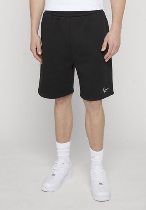Homme portant un short décontracté noir avec une taille élastique, des chaussettes blanches et des baskets blanches, debout devant un fond clair uni.