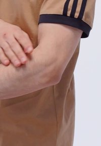 Camisa marrón de manga corta con cuello negro; el brazo está ligeramente tonificado con textura de piel visible y pequeñas imperfecciones.