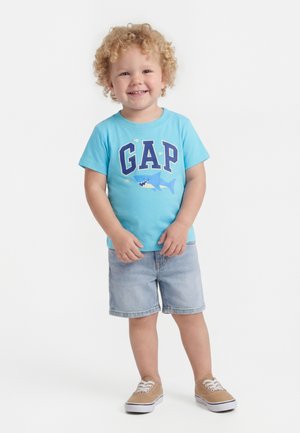 Copil mic cu păr blond creț, purtând un tricou albastru GAP cu un grafic cu rechin, pantaloni scurți din denim și adidași bej, zâmbind.