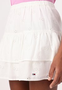 White cotton skirt with layered ruffles, featuring embroidered floral patterns and a small logo tag in red, white, and blue.