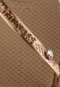 Close-up of a bronze-colored sandal strap embossed with "havaianas" and decorated with a round iridescent gem.
