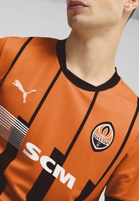 Camiseta atlética naranja con rayas negras, cuello negro, logo de Puma en el pecho, presenta un emblema del Shakhtar Donetsk y texto blanco "SCM".