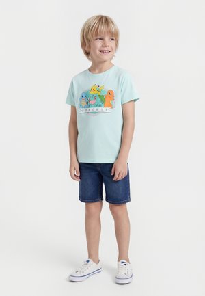 Lyshåret dreng iført lyseblå Pokémon t-shirt, mørke denimshorts og hvide sneakers, stående og kiggende til siden med et smil.