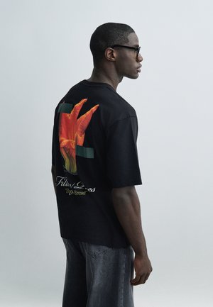 Férfi fekete szemüvegben, fekete pólóban, amelyen nagy piros virág grafika és szöveg található a háton, valamint kifakult kék farmerben, jobbra néz egy egyszerű háttér előtt.