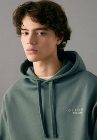 Sudadera con capucha verde con bolsillo frontal, cordones y texto bordado "CHILLIN' IN THE LOBBY" en color beige claro. Tejido suave y texturizado.