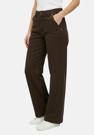 Persona che indossa pantaloni larghi marrone scuro e sneakers bianche, in piedi con una mano in tasca contro uno sfondo semplice.