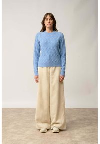 Jasnoniebieski sweter dziany z teksturowym wzorem, w zestawieniu z kremowymi spodniami o szerokich nogawkach z prążkowanego materiału. Model stoi na neutralnym tle.