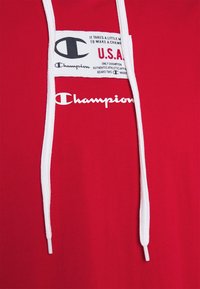 Punainen huppari, joka on valmistettu puuvillasta ja jossa on valkoiset kiristysnauhat. Edessä keskellä on Champion-logo ja -etiketti, jossa on teksti.