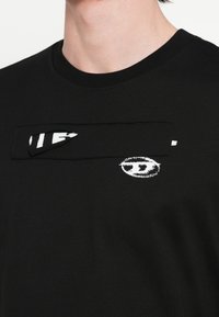 Μαύρο βαμβακερό T-shirt με στρογγυλή λαιμόκοψη, που διαθέτει υφασμάτινο ύφασμα με υφή και λευκά τυπωμένα σχέδια, συμπεριλαμβανομένου λογοτύπου και στυλιζαρισμένου κειμένου.