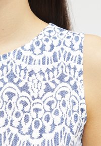 Valkoinen tekstiili, jossa on monimutkainen sininen pitsikuvio, jossa on kukkakuvioita ja geometrisia muotoja. Suorastaan sileä rakenne, pyöreä kaula-aukko ja olkaimeton tyyli.