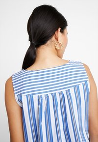 Top senza maniche blu e bianco a righe con dettaglio arricciato sul retro, tessuto liscio e scollatura rotonda. Orecchini a cerchio dorati visibili.