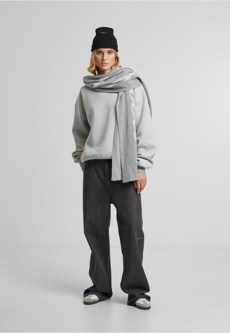Grå oversized sweatshirt, mørke brede jeans, grå tørklæde og sort strikket hue. Hvid tekstmønster på tørklædet, glat tekstur, afslappet stil.
