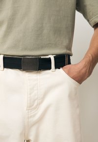 Cintura nera intrecciata con fibbia rettangolare metallica, indossata sopra pantaloni bianchi. La maglietta è di colore verde chiaro, a maniche corte.