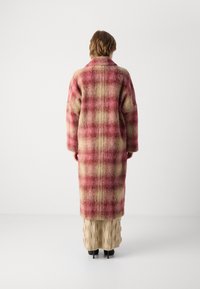 Geruite wollen jas in roze- en beigetinten, lange snit, oversized pasvorm, brede kraag, zichtbare naden, gedragen over een gestructureerde beige rok.