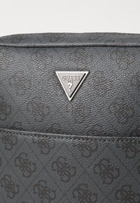 Bolso de material texturizado negro con un sutil patrón en relieve y un logo de marca triangular plateado con el texto "GUESS."