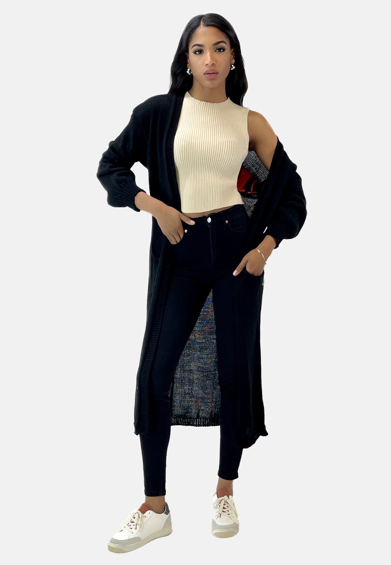 Czarny dzianinowy kardigan na beżowym, prążkowanym topie bez rękawów, zestawiony z ciemnymi jeansami i białymi trampkami. Posiada długi, otwarty krój z kieszeniami.
