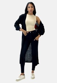 Svart lång cardigan över en beige ribbad topp, kombinerad med svarta jeans och vita sneakers. Funktioner inkluderar sidslitsar i cardiganen.