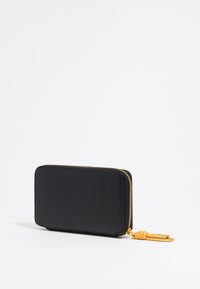 Cartera de cuero negra con un acabado texturizado, forma rectangular, cierre dorado y detalle de borlas naranjas. Cerrada, mostrando solo un lado.