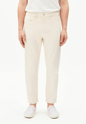 Man wearing off-white jeans and white slip-on shoes standing against a plain white background.