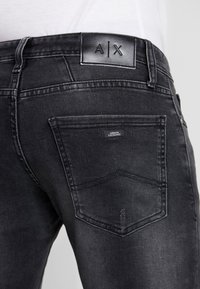 Svarta denimjeans med en lädermidjeetikett med "A|X." Bakfickan har en liten etikett med "Armani Exchange." Slät, något blekt textur.