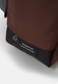 Hnědá a černá nylonová taška s hladkým povrchem, s visačkou, která zní: "Tato taška byla odpad! Je vyrobena z recyklovaného nylonu."