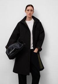 Cappotto oversized nero con colletto alto, foderato in verde e dotato di due tasche frontali. Il modello tiene in mano una borsa nera lucida. Sotto indossa una maglietta bianca.