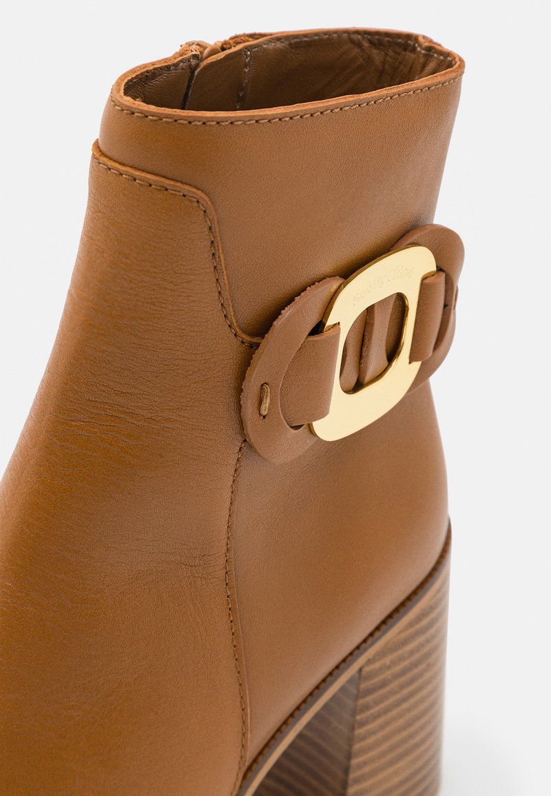 Bottine en cuir marron avec une surface texturée, une accentuation en boucle dorée et un talon en bois empilé. Fini lisse et fermeture éclair.