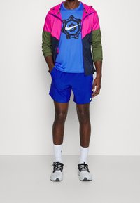 Hombre con una chaqueta rosa, oliva y negra sobre una camiseta de logo de Nike azul, pantalones cortos azules, calcetines blancos y zapatillas blancas y negras.