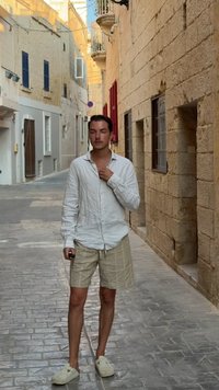 Chemise en lin à manches longues, shorts beige clair avec un motif à carreaux, et chaussures à enfiler beiges, se tenant dans une ruelle en pierre avec des volets fermés.