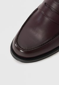 Mocassin en cuir bordeaux foncé avec une finition lisse, un bout arrondi et des surpiqûres décoratives le long du chausson, reposant sur une semelle en caoutchouc noir.