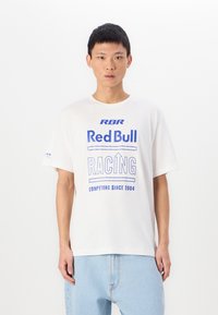 T-shirt en coton blanc avec un texte imprimé bleu : "RBR Red Bull RACING COMPETING SINCE 2004." Coupe décontractée avec des manches courtes. Jean en denim léger.