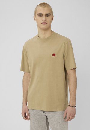 Jeune homme aux cheveux blonds courts portant un t-shirt beige avec un petit logo crabe rouge sur le côté gauche de la poitrine et un jean gris clair.