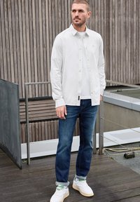 Bílá košile s dlouhým rukávem na knoflíky přes bílém tričku, v kombinaci s tmavě modrými džíny a bílými teniskami. Hladká tkanina a uvolněný střih.