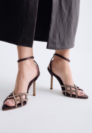 Pies con sandalias de tacón alto marrón oscuro con tacones decorados con cuentas y tiras cruzadas, combinadas con pantalones oscuros hasta el tobillo.